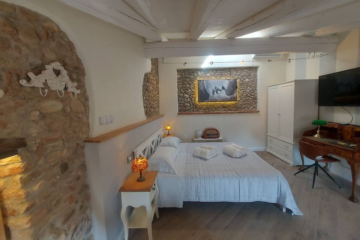 A bedroom with a stone wall, a bed with white bedding, a bedside table with a lamp, a TV on the wall, and a desk with a chair.