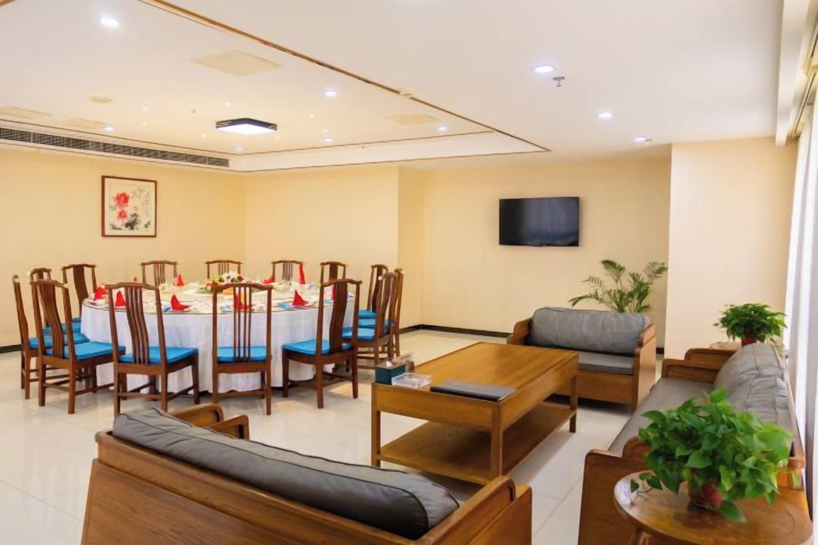 A hotel room with a dining table set for four, a sofa, a TV mounted on the wall, and a potted plant.