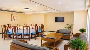 A hotel room with a dining table set for four, a sofa, a TV mounted on the wall, and a potted plant.