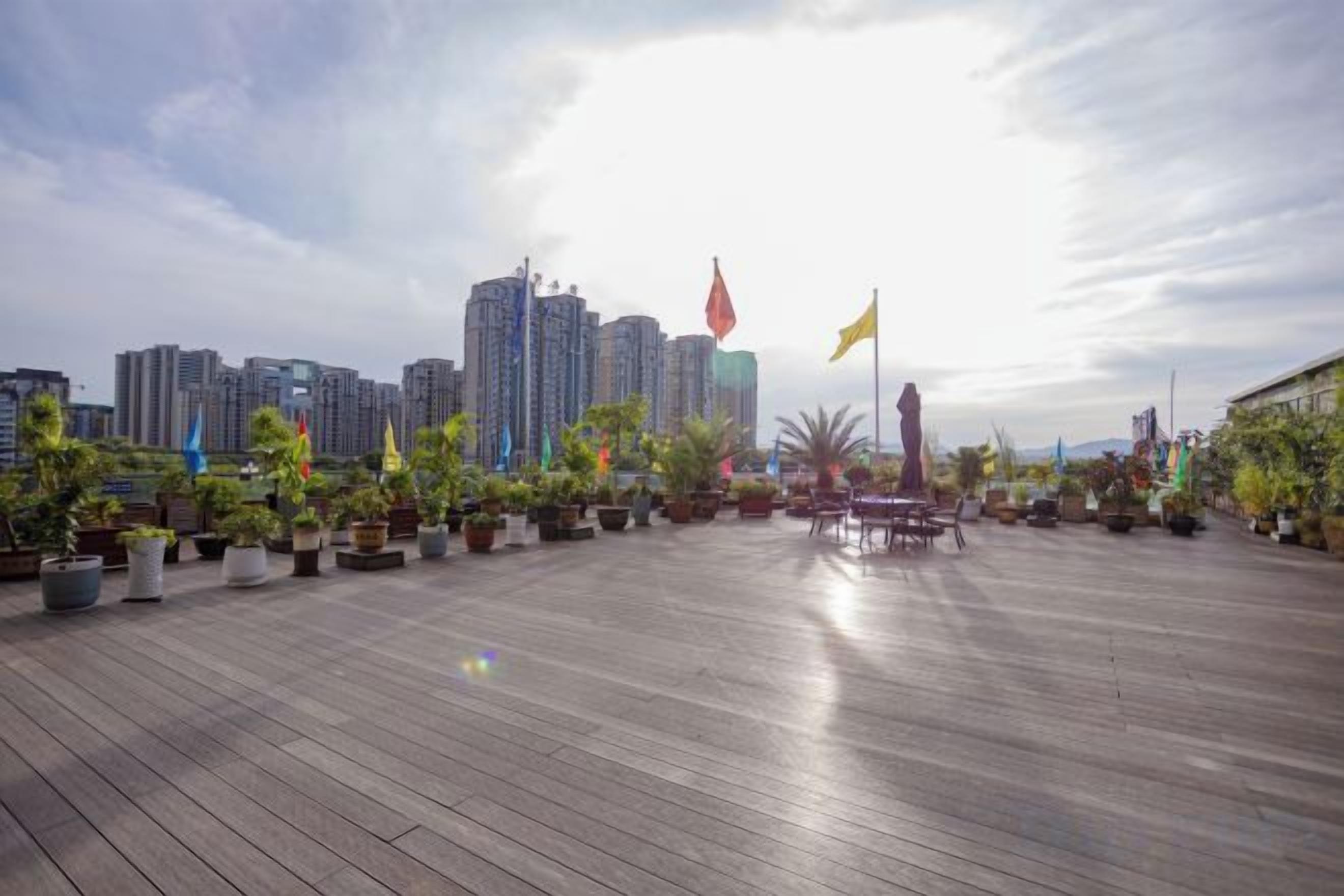 Una terraza de madera al aire libre con plantas en macetas y banderas, con vista a un paisaje urbano con rascacielos.