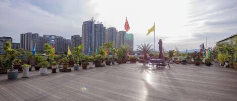 Una terraza de madera al aire libre con plantas en macetas y banderas, con vista a un paisaje urbano con rascacielos.