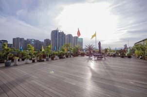 Una terraza de madera al aire libre con plantas en macetas y banderas, con vista a un paisaje urbano con rascacielos.