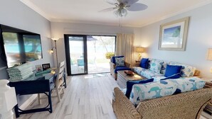 A living room with a sofa, a coffee table, a TV, and a desk. There is a door leading to an outdoor area. - Awesome Oceanfront Grd Flr Condo on Spectacular 7 Mile Beach – #19 TIC (Grand Cayman)