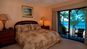 Une chambre à coucher avec un lit, deux tables de chevet avec des lampes, un tableau au mur et un balcon avec des chaises donnant sur un paysage.