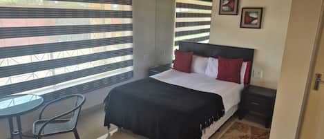Quarto com cama, mesa de cabeceira, cadeira e janela com persianas.