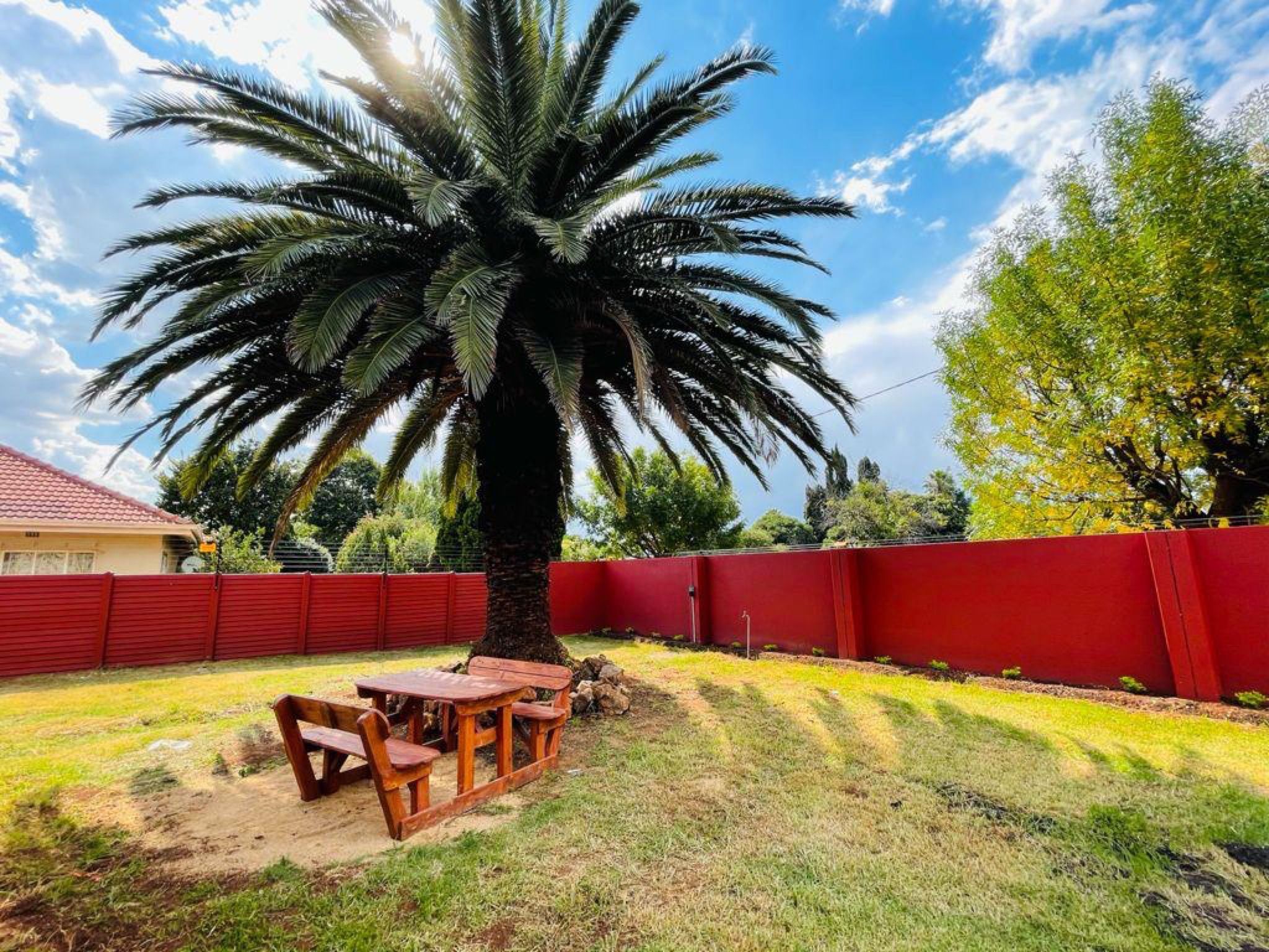 A backyard with a wooden table and chairs, a palm tree, and a red fence.