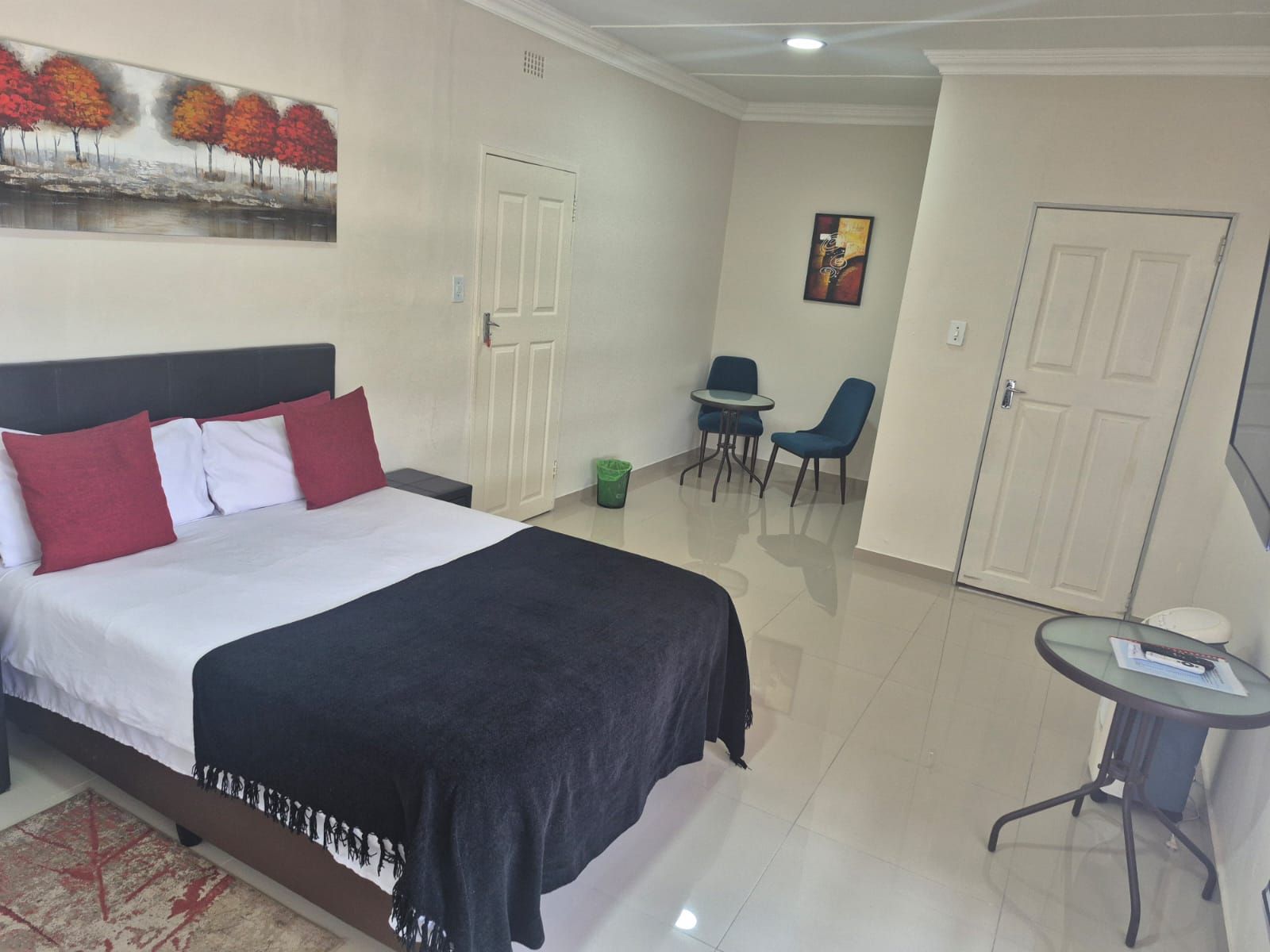Quarto de hotel com cama, uma pequena mesa com cadeiras, um quadro na parede e uma porta.