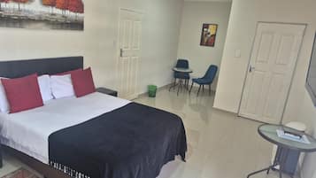 Quarto de hotel com cama, uma pequena mesa com cadeiras, um quadro na parede e uma porta.