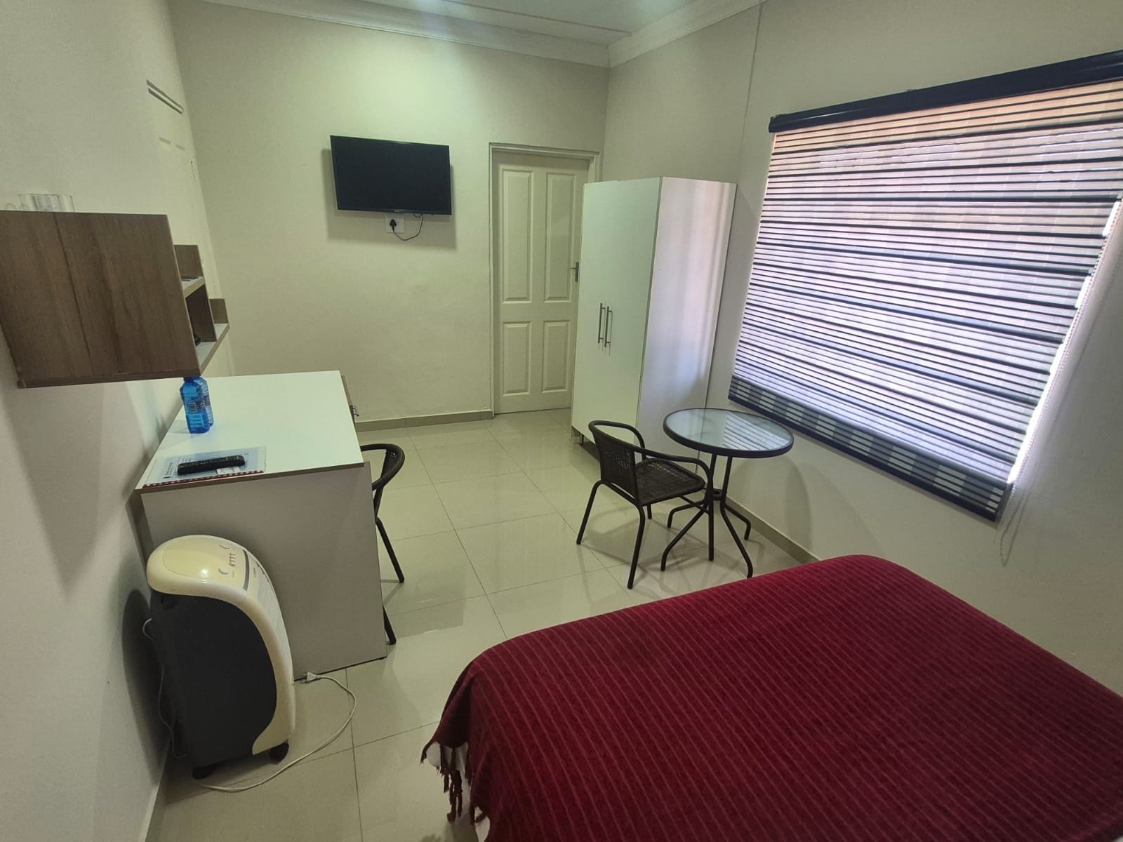 A small, clean room with a bed, a desk, a chair, a TV, and a window with blinds.