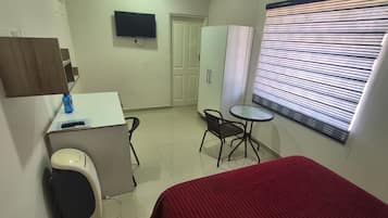 A small, clean room with a bed, a desk, a chair, a TV, and a window with blinds.