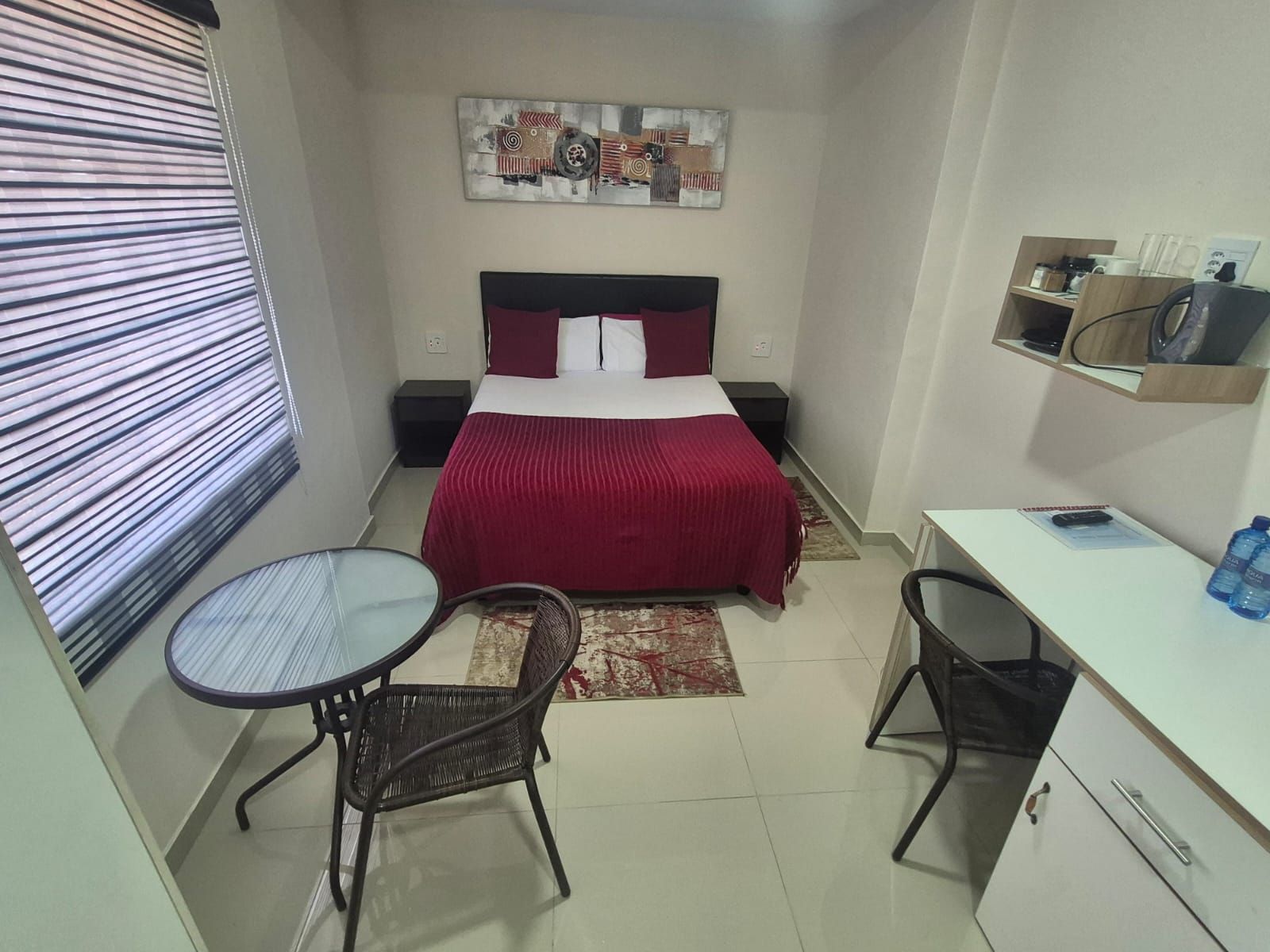 Une chambre d’hôtel de taille réduite, comprenant un lit, un bureau, deux chaises, une petite étagère avec quelques objets et une fenêtre avec des stores.