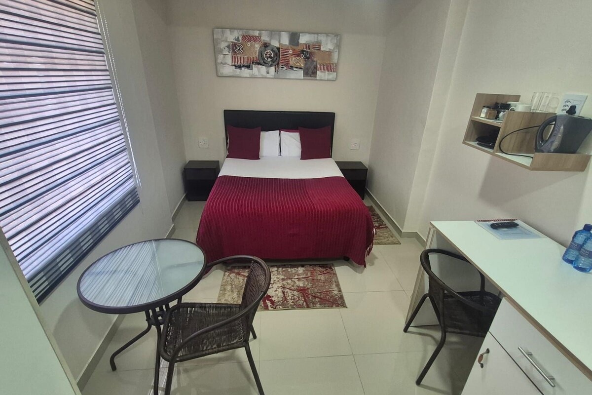 Quarto de hotel compacto com cama, escrivaninha, duas cadeiras, uma prateleira com objetos e uma janela com persianas.