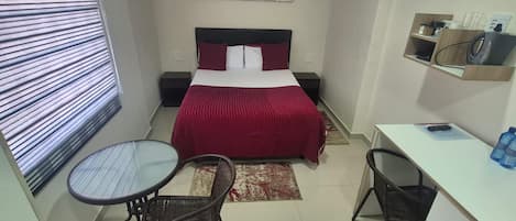 A compact hotel room with a bed, a desk, two chairs, a small shelf with items, and a window with blinds.