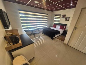 Ein modernes Hotelzimmer mit einem Bett, einem kleinen Schreibtisch, einem Stuhl und einem Fenster mit Jalousien.
