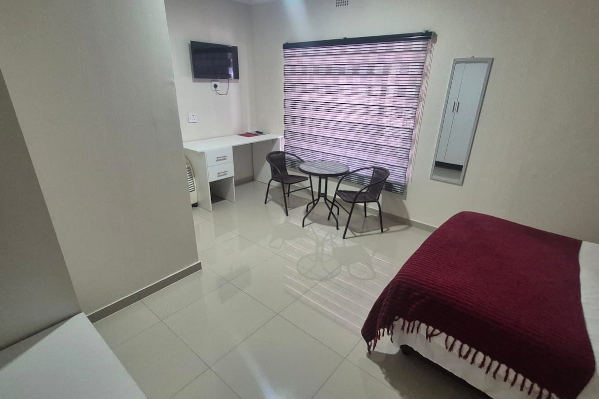 Une chambre à coucher comprenant un lit, un bureau, deux chaises, un téléviseur et une fenêtre avec des stores.