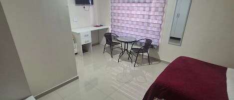 A bedroom with a bed, a desk, two chairs, a TV, and a window with blinds.