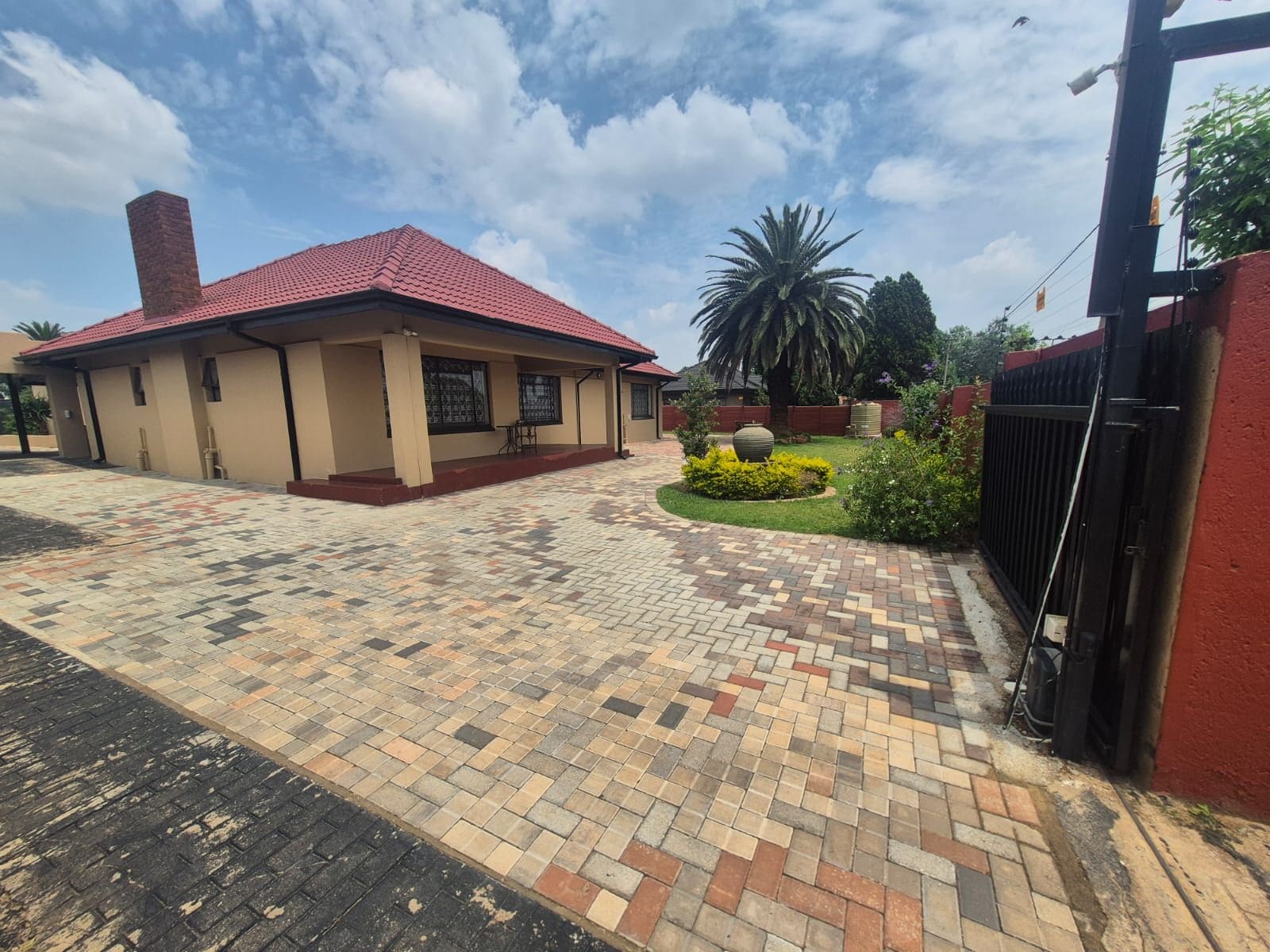 A building with a red roof, a paved entrance area, and a garden with a palm tree.