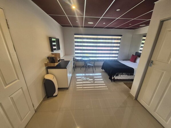 A compact hotel room with a kitchenette, a bed with a black headboard, a small table and chairs, and a large window with blinds.