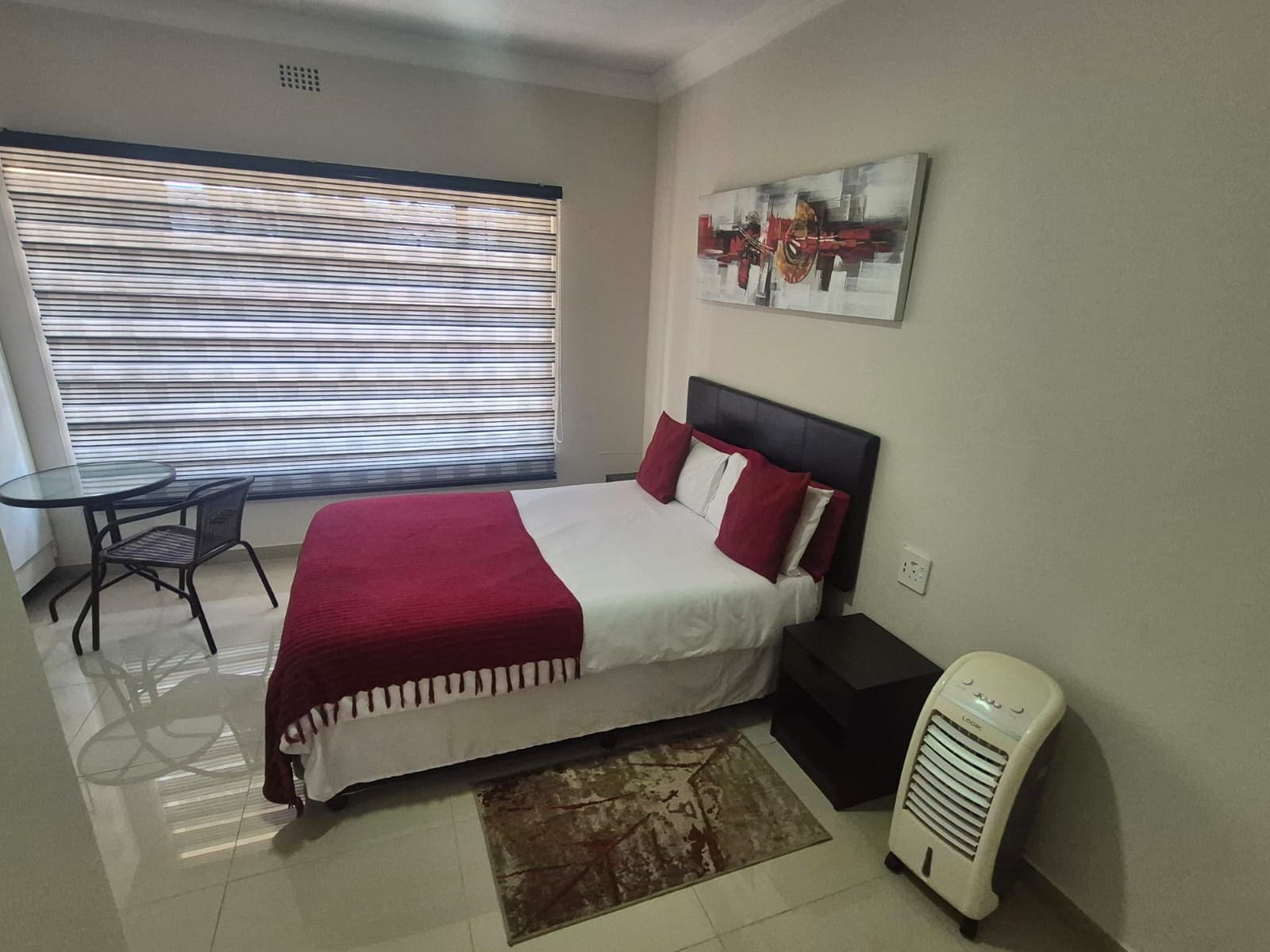 A hotel room with a bed, a small table, a chair, a window with blinds, a radiator, and a painting on the wall.