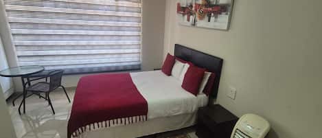 Una camera d'albergo con un letto, un tavolino, una sedia, una finestra con persiane, un termosifone e un quadro appeso al muro.