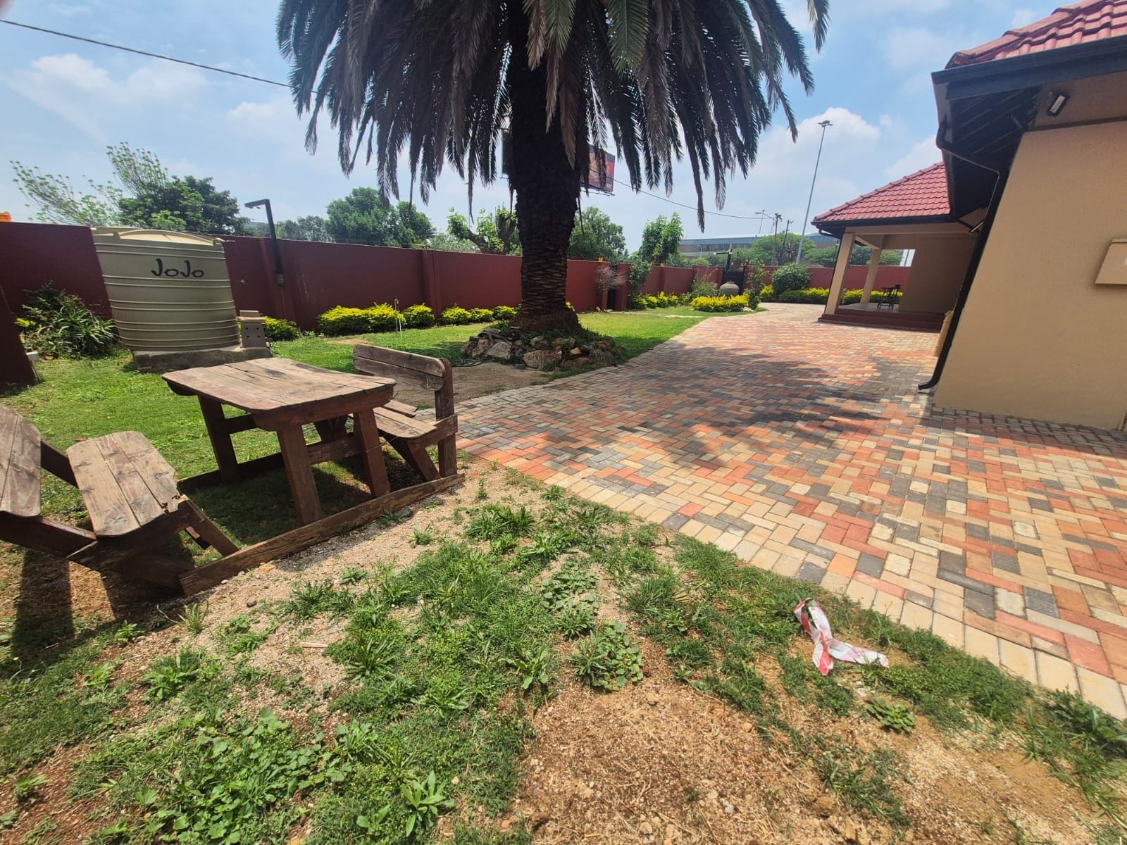 Uma área de pátio com mesa de piquenique e bancos, cercada por um caminho de tijolos e uma palmeira.