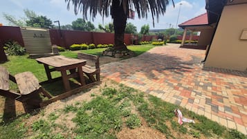 Uma área de pátio com mesa de piquenique e bancos, cercada por um caminho de tijolos e uma palmeira.