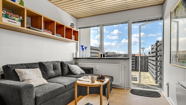 A modern living room with a grey sofa, wooden shelves, and a large window with a view of a fenced outdoor area. - Amazing apartment in Ebeltoft with WiFi (Ebeltoft)