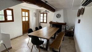 A dining area with a long wooden table, black chairs, and a bench. There are two windows with curtains, a stone fireplace, and a wall-mounted clock. - La Bastide des Oliviers (COTIGNAC)