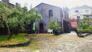 A courtyard with a stone pathway leading to a building with a dark facade and a small window. There are potted plants and a white chair near the entrance. - A True Sicilian guesthouse with Pool on Mt. Etna, perfect for Families & Groups. (Puntalazzo)