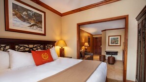 Une chambre d’hôtel avec un grand lit, une tête de lit en bois, un tableau au mur, un bureau avec une lampe et une porte donnant sur une autre pièce.