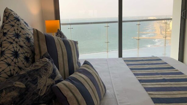 Ein Schlafzimmer mit großem Fenster und Blick auf die Küste, mit einem Bett mit gestreifter Bettwäsche sowie einer Brücke und einem Strand.