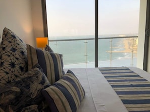 A bedroom with a large window overlooking a coastal view, featuring a bed with striped bedding and a view of a bridge and beach.