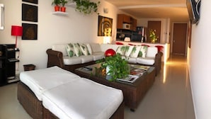 Ein Wohnzimmer mit einer Sofagarnitur, einem Couchtisch mit Zeitschriften, einem an der Wand befestigten Fernseher und einer Küchenzeile im Hintergrund.