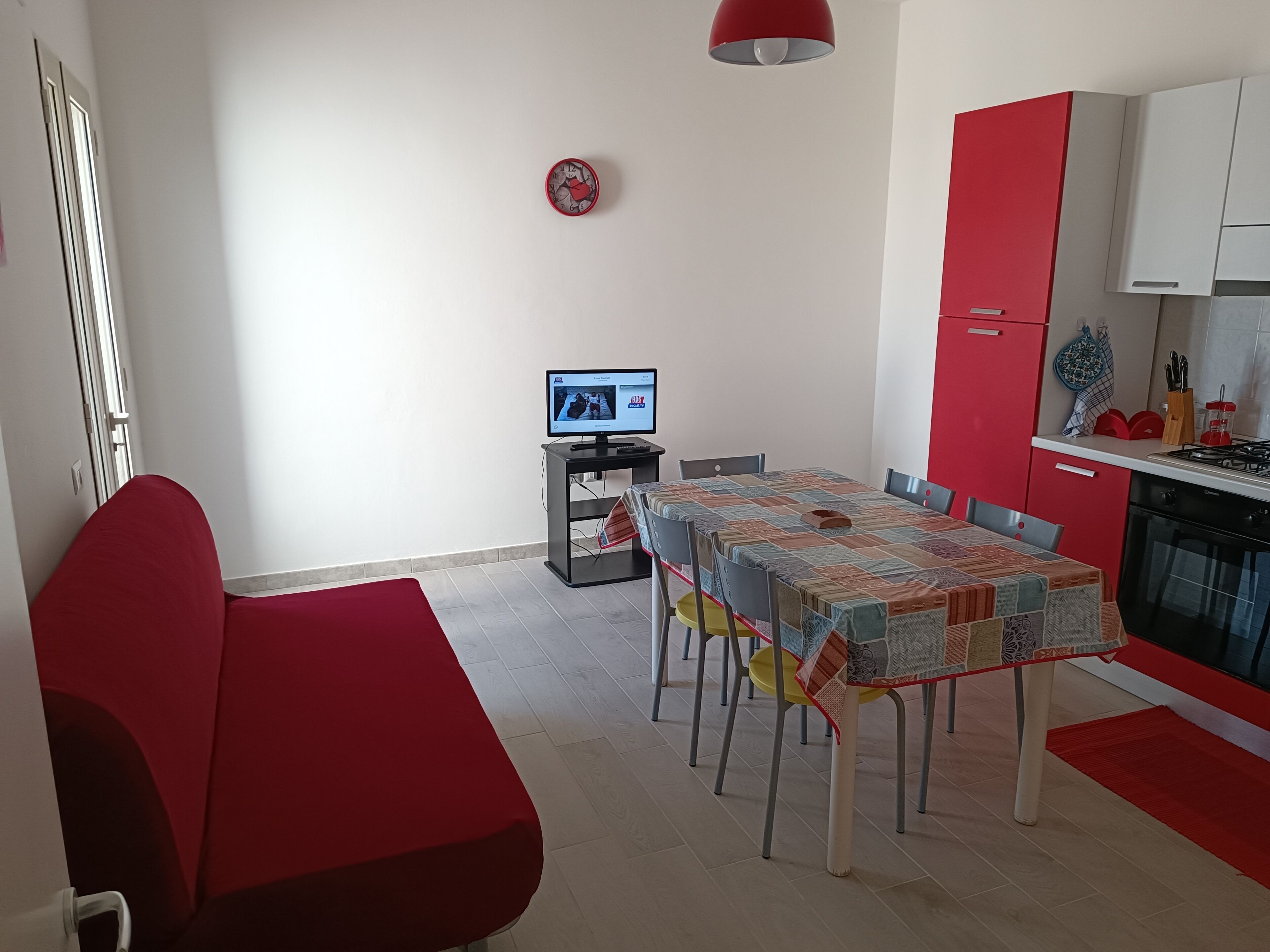 Eine moderne Küche mit roten Schränken, einem Esstisch mit Stühlen, einem Fernseher auf einem Schrank und einem roten Sofa.