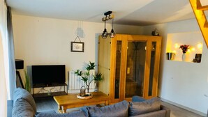 Un salon avec un canapé gris, une table basse en bois, un téléviseur posé sur un meuble, une plante et un buffet en bois doté de portes en verre.
