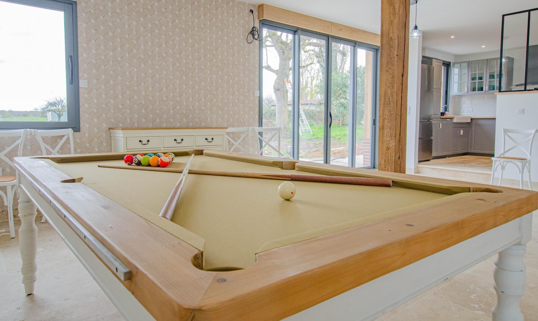Une table de billard avec des billes, une table en bois et une fenêtre donnant sur un jardin.
