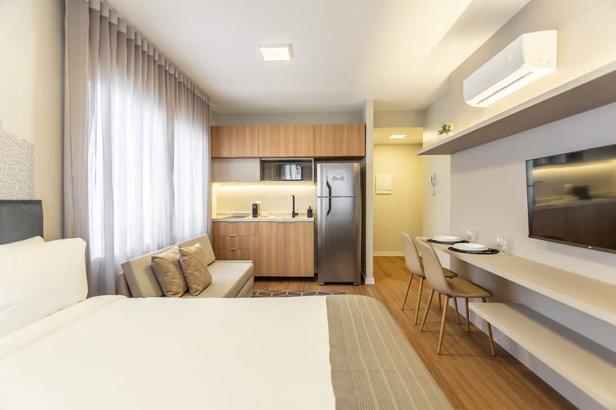 A modern hotel room with a bed, a sofa, a dining area, and a kitchenette.