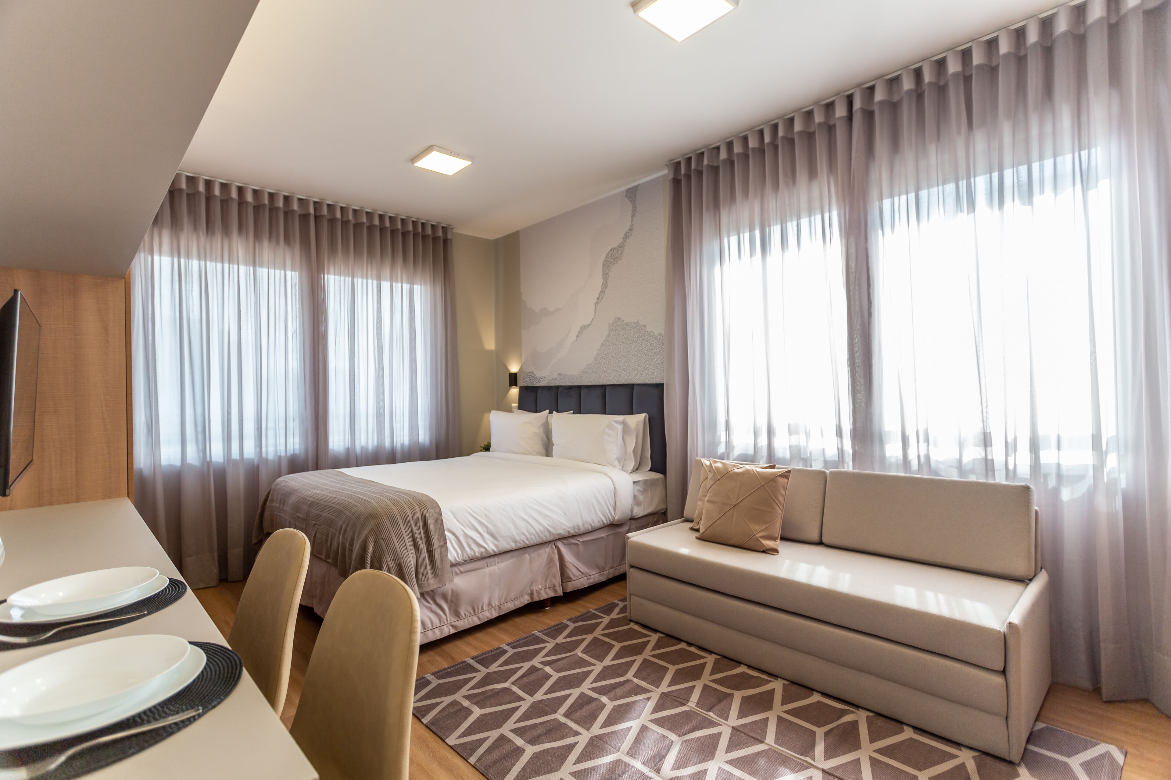 Ein modernes Hotelzimmer mit Bett, Sofa, Esstisch und Vorhängen.