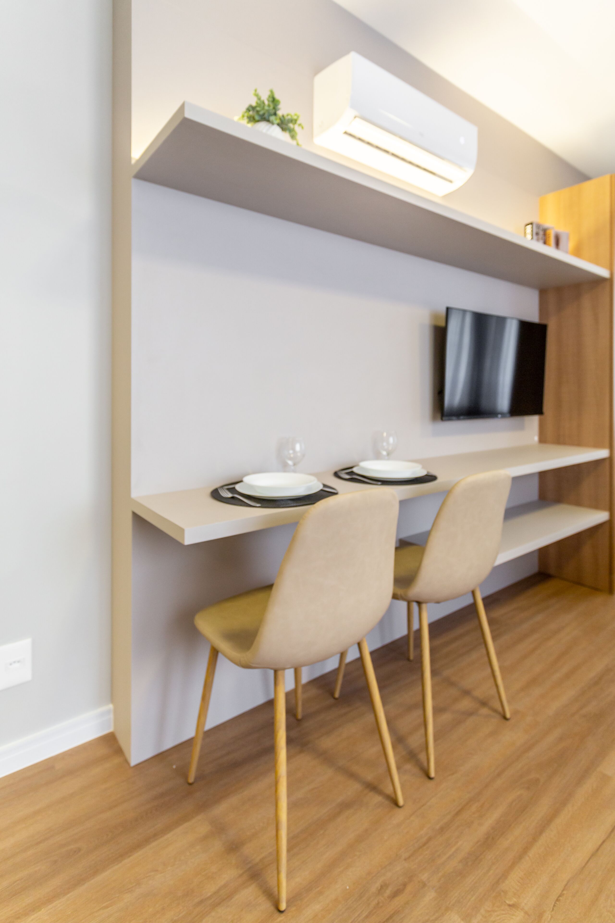 Un comedor moderno con suelo de madera, una mesa de comedor blanca, dos sillas, un televisor de pantalla plana montado en la pared y un pequeño estante con una planta.