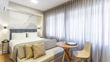 A hotel room with a bed, a small round table with a chair, a nightstand with a lamp, and a large window with curtains.