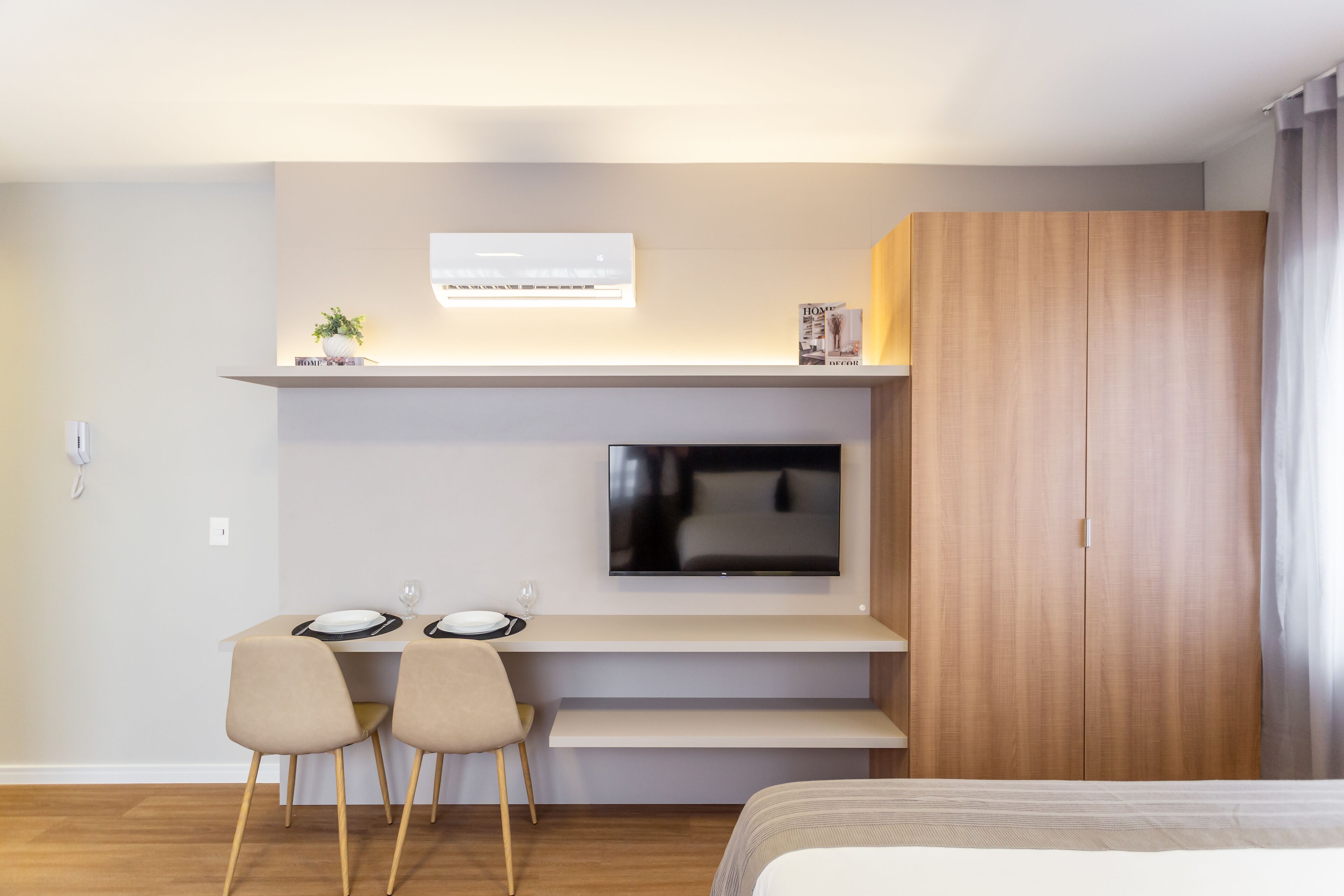 A modern hotel room with a flat-screen TV mounted above a desk, two chairs, a bed, and a wooden wardrobe.