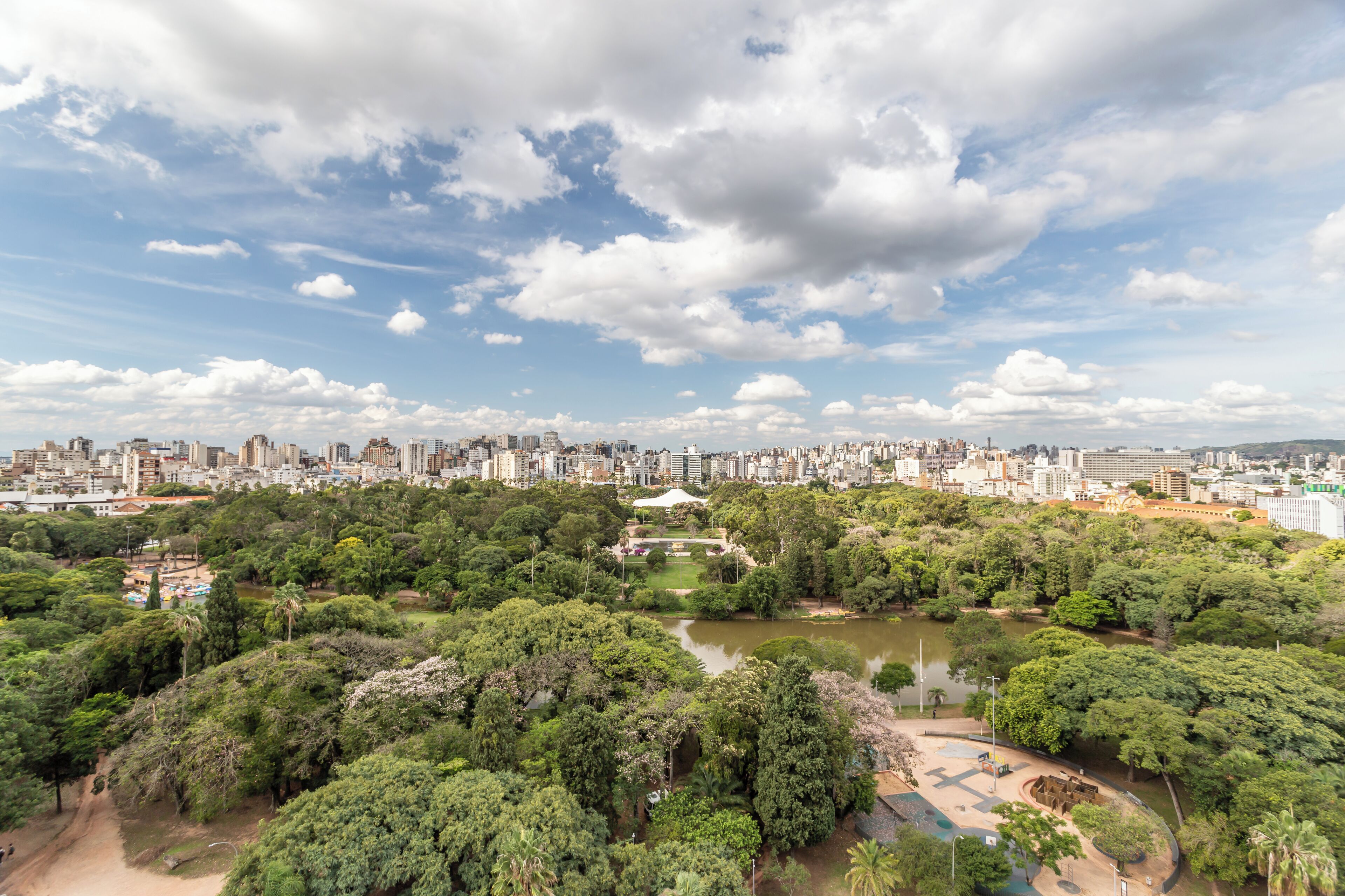 Un parque con un lago, rodeado de árboles densos y un horizonte urbano al fondo.