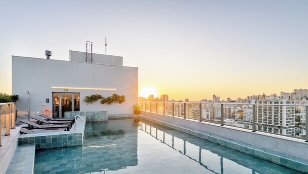 Una piscina en la azotea con vistas a la ciudad al atardecer, un edificio blanco con una puerta y plantas, y una barandilla.
