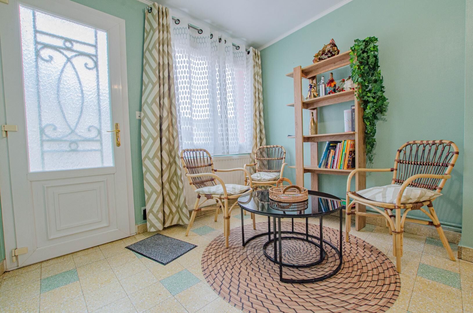 Una habitación con una puerta blanca, dos sillas de mimbre, una mesa redonda, una estantería y una planta en maceta.