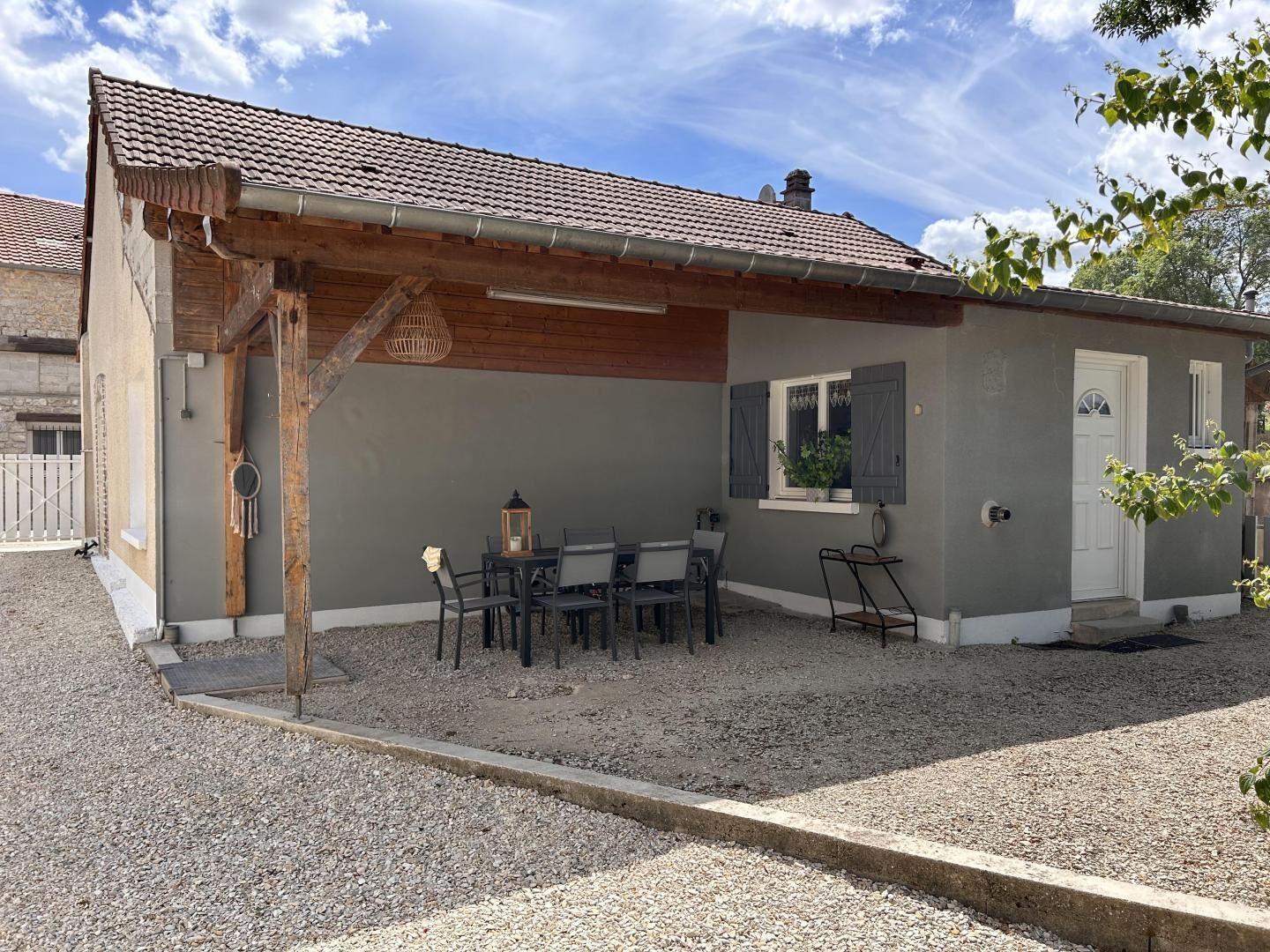 A small building with a tiled roof, a wooden pergola, and a gravel patio with outdoor seating.
