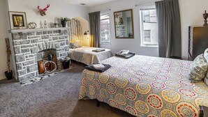 Un dormitorio con chimenea de piedra, una cama con colcha estampada, una mesita de noche, un aparador y una ventana con cortinas.