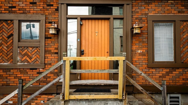 Un edificio de ladrillo con puerta de madera marcada con el número 7, una escalera de madera y dos lámparas de pared tipo farol.