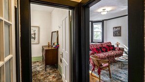Une pièce avec un tapis à motifs, un canapé recouvert d’un tissu rouge et noir à motifs, une chaise en osier et une petite table avec une lampe.