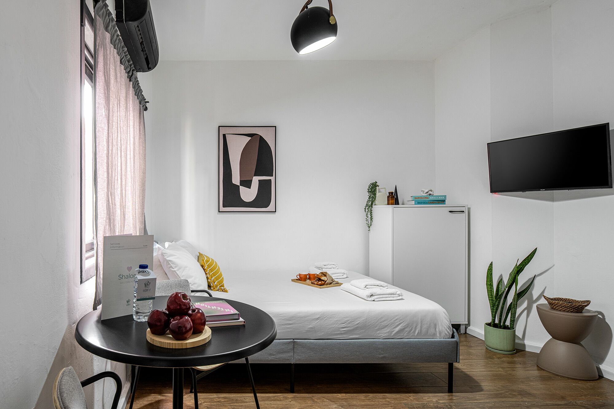 Habitación de hotel moderna con una cama, una mesita con manzanas y un libro, un televisor y un refrigerador.