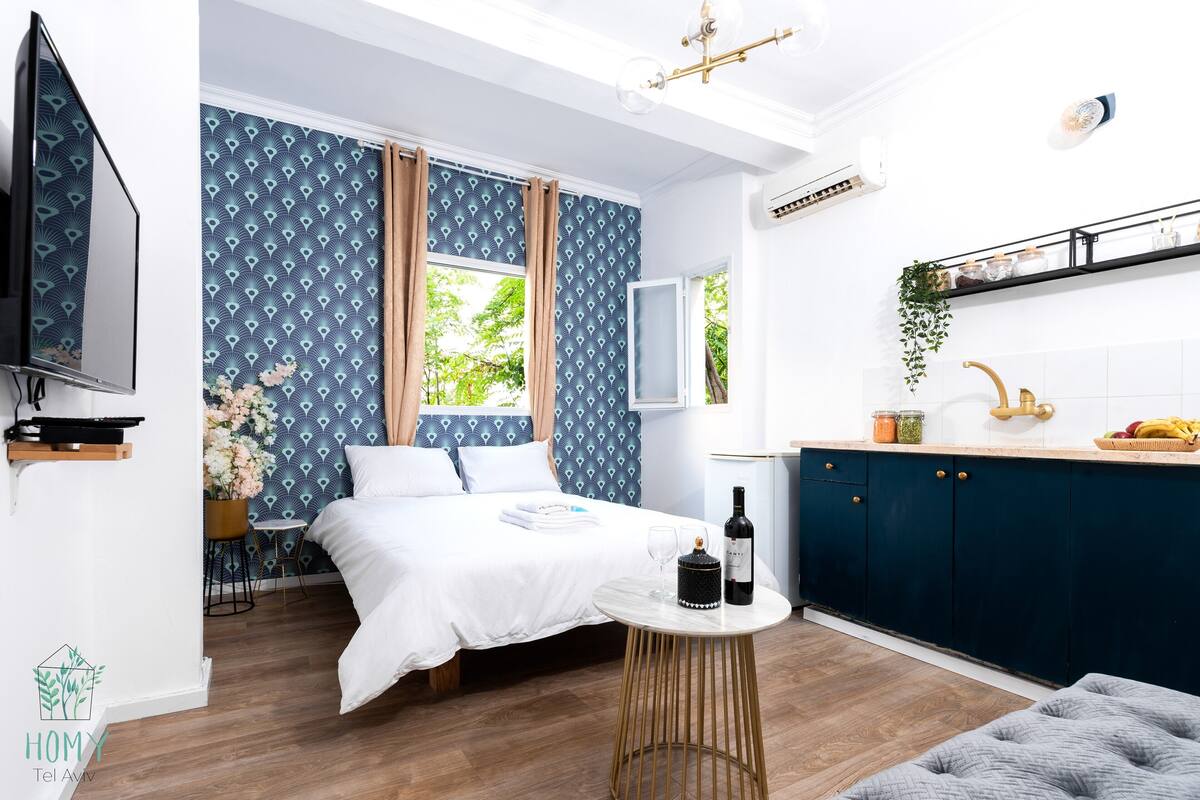 Una habitación de hotel moderna con una cama, una pequeña mesa redonda con una botella y vasos, un televisor, una zona de cocina con un lavamanos y armarios, y una ventana con cortinas.